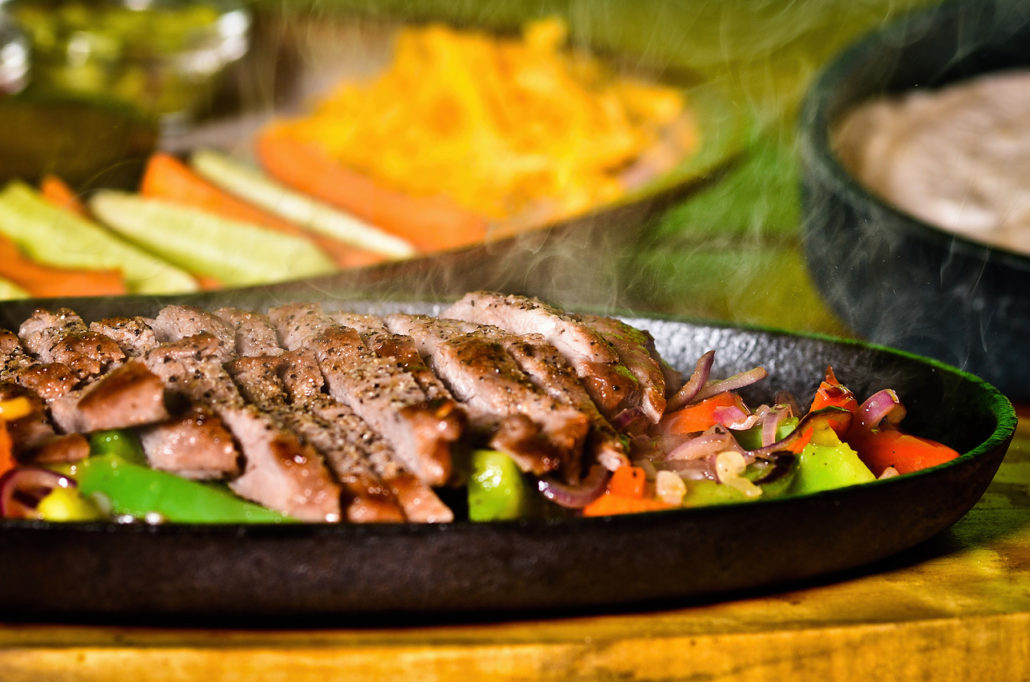 Fajitas na rozpálené pánvi, v pozadí strouhaný sýr a domácí salsa